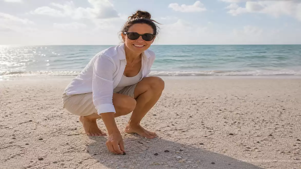 A woman wearing sunglasses and a white shirt kneels on the beach in front of the sea.