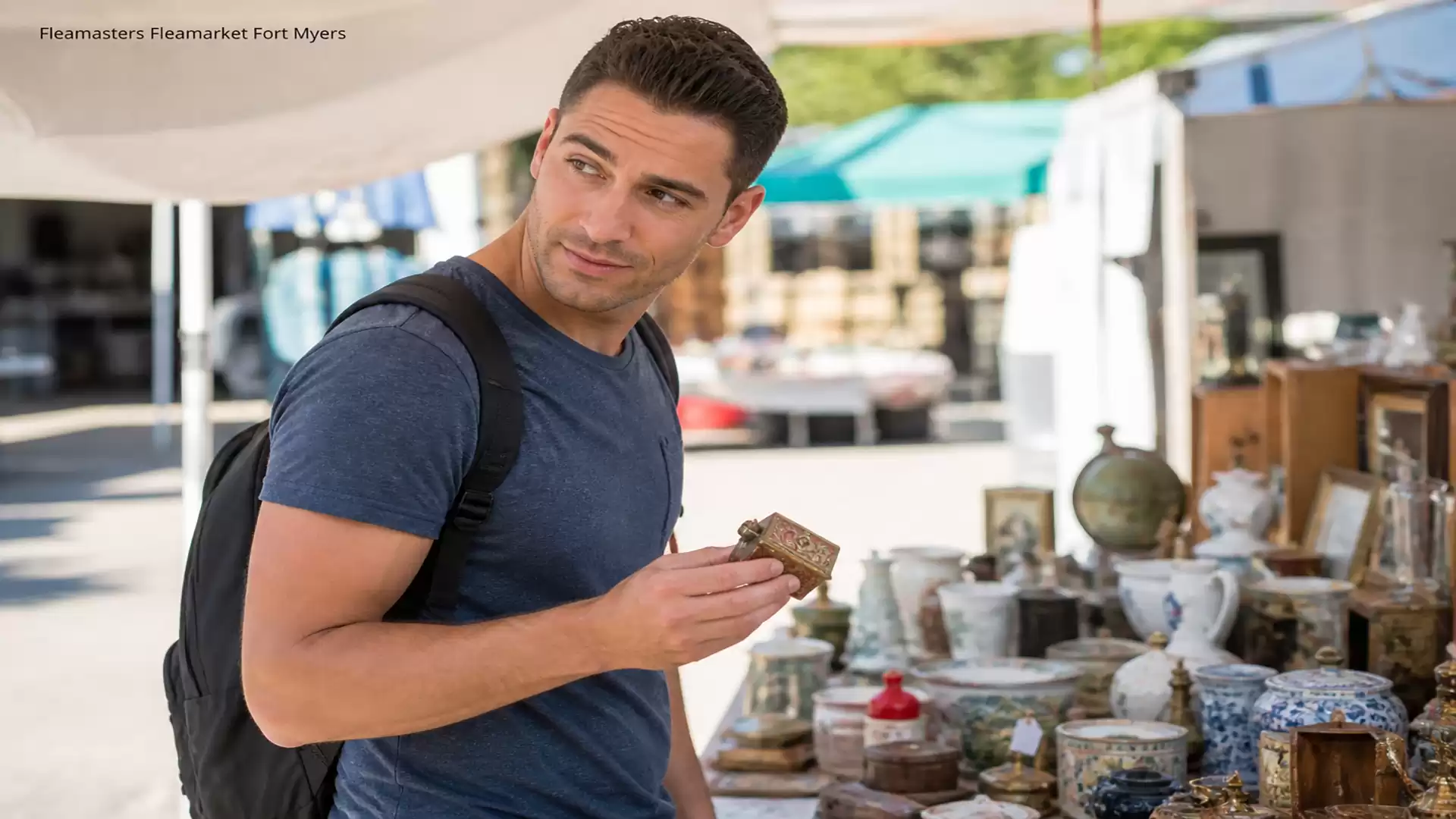 A man with dark hair and a blue T-shirt browsing antiques at a bustling flea market with many stalls.