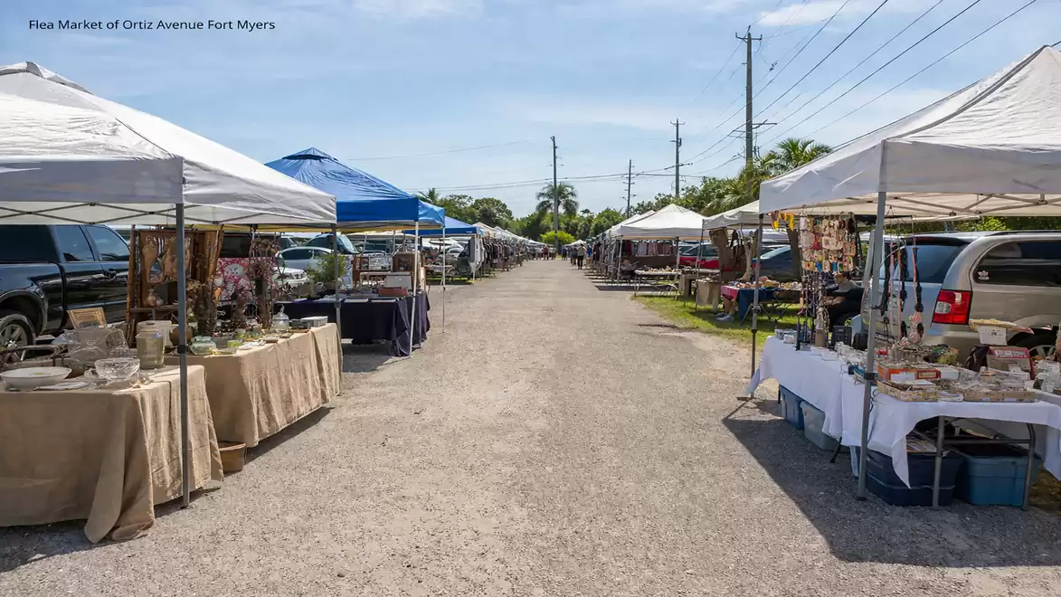 A vibrant flea market on a sunny day featuring various vendor stalls and white canopies on both sides of a gravel path.