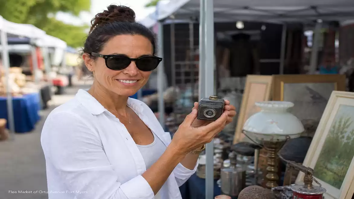 A woman wearing sunglasses holds an antique camera at a flea market stall and smiles.