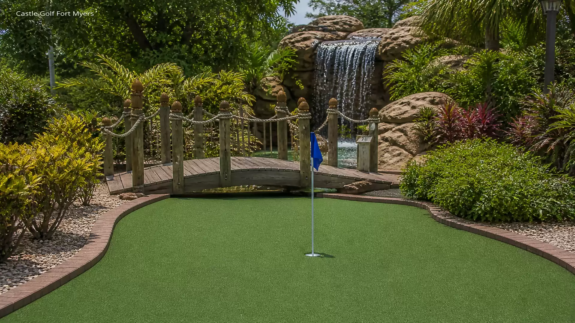 An attractive mini-golf course featuring a wooden bridge and a waterfall in the background, surrounded by lush vegetation and well-maintained plants.
