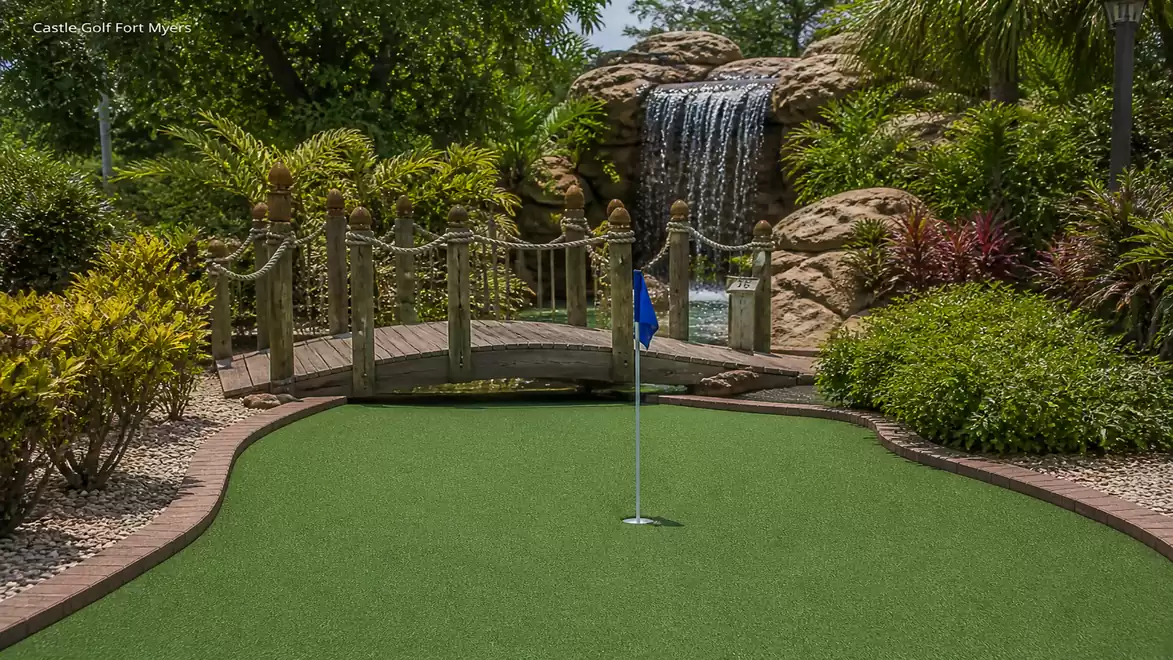 An attractive mini-golf course featuring a wooden bridge and a waterfall in the background, surrounded by lush vegetation and well-maintained plants.