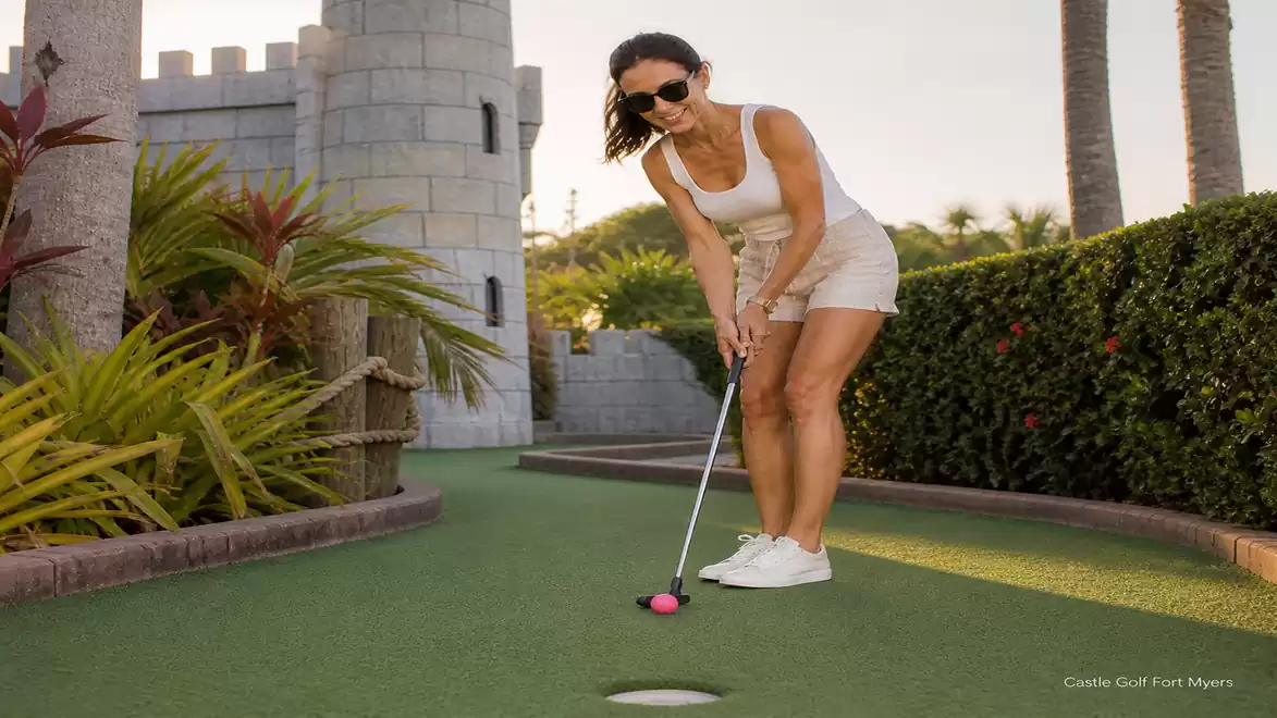 Woman playing mini-golf on a green course with a castle in the background and palm trees at sunset.