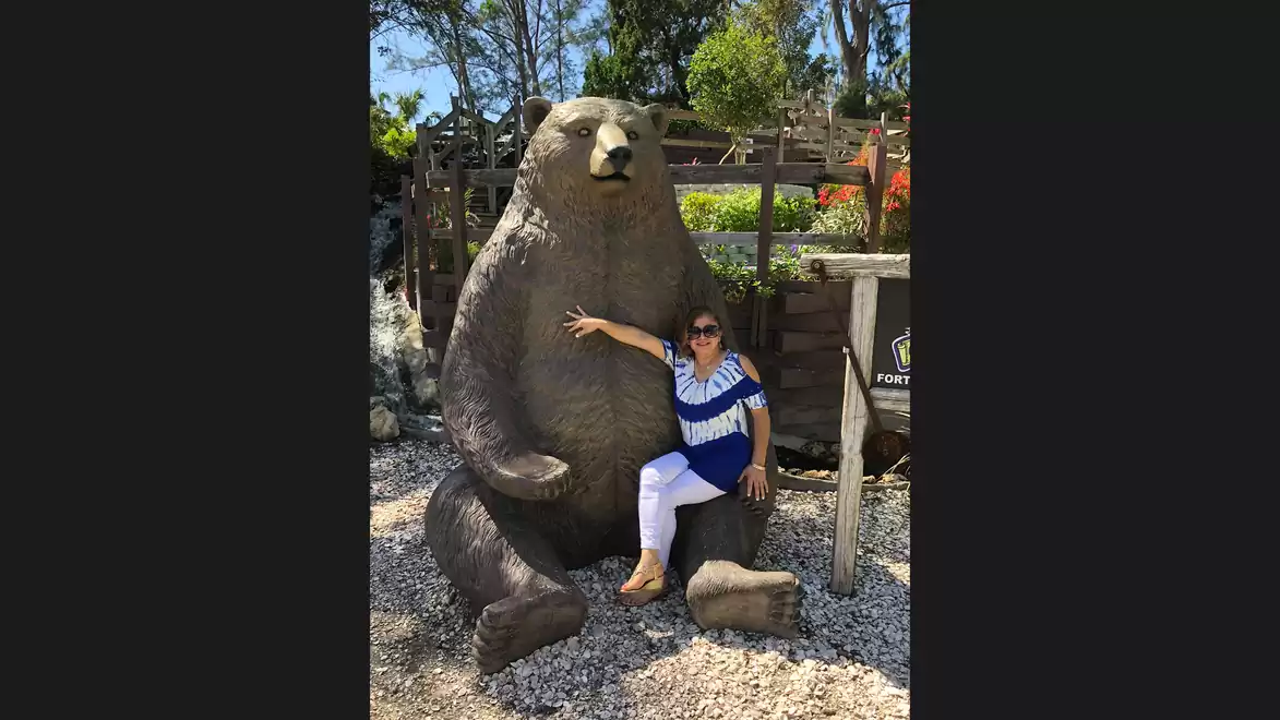 Woman posing in front of a large bear statue outdoors, surrounded by trees and gravel.