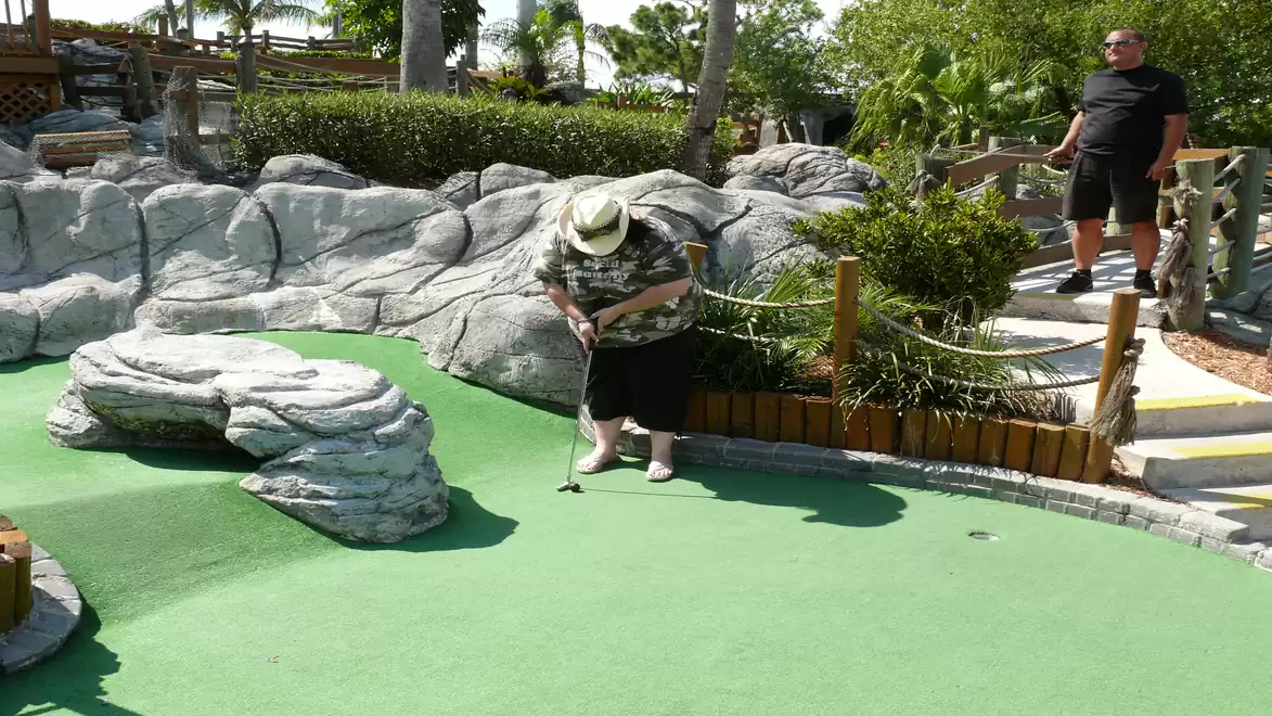 An elderly man playing mini-golf on a green course surrounded by rocky decorations, while another man watches.