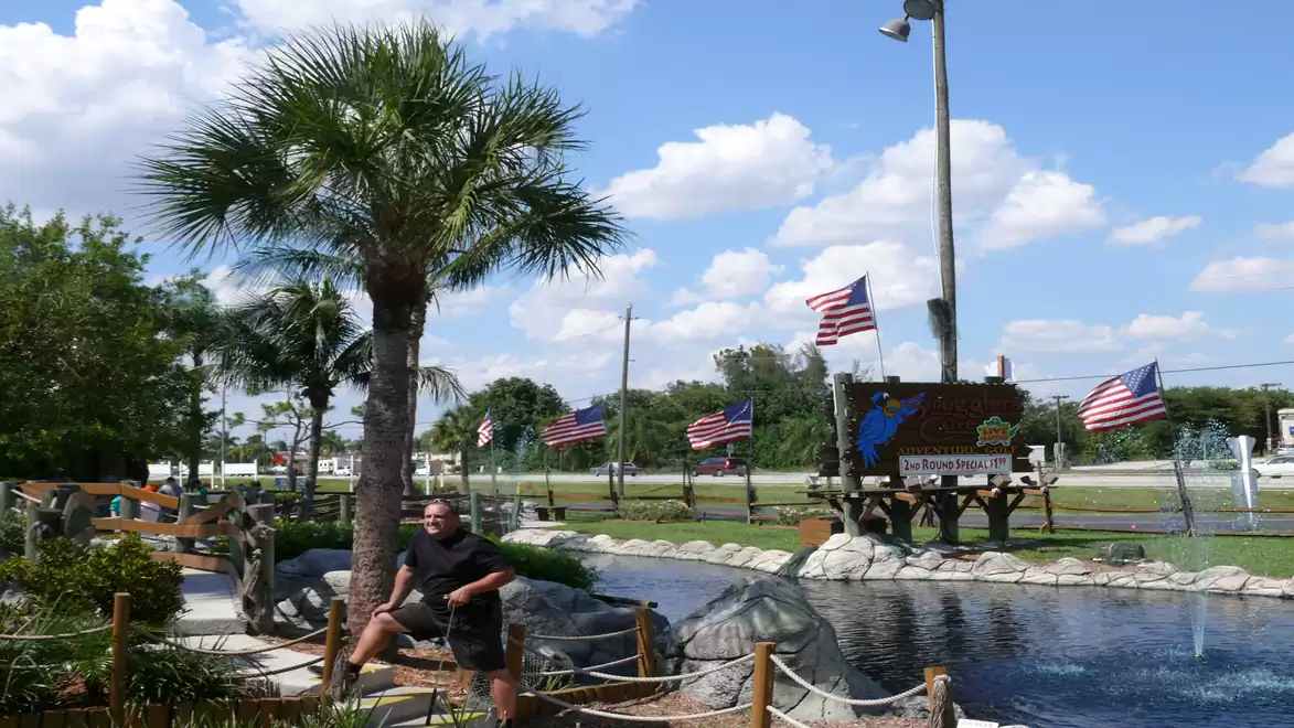 A man sits on a stone railing next to an artificial pond, surrounded by palm trees and waving American flags under a clear blue sky.