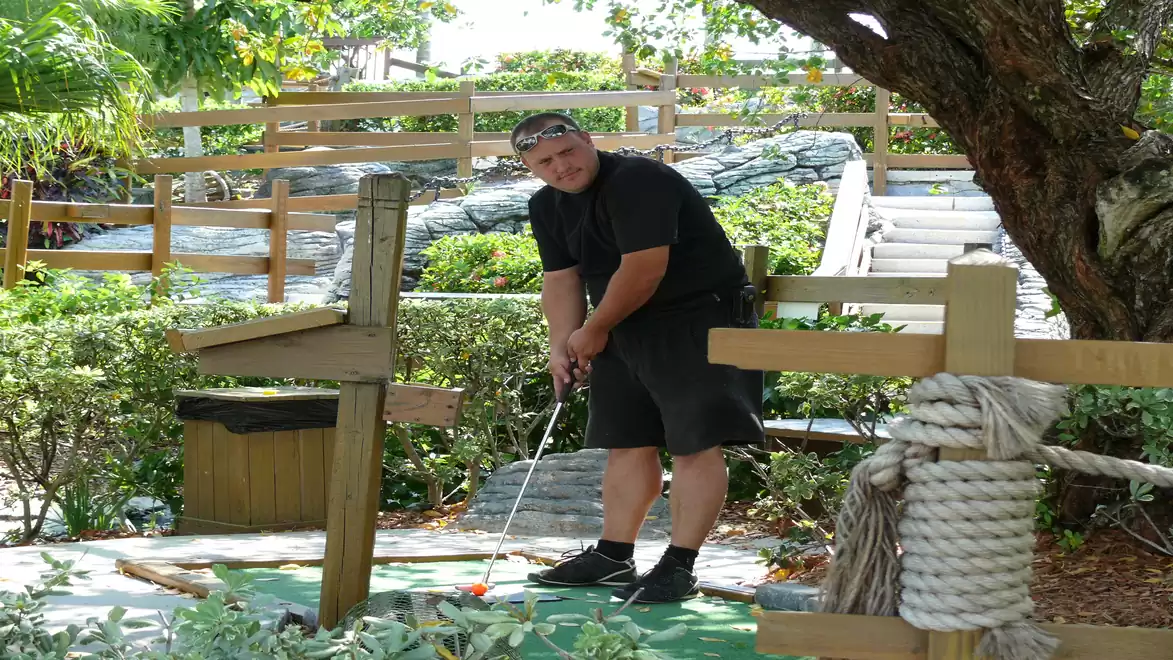 Man playing mini-golf on a green outdoor course, surrounded by trees and wooden railings.