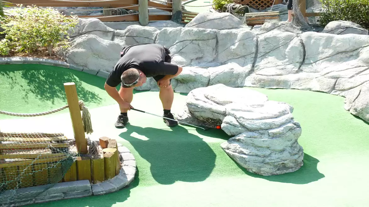 Man playing mini golf, bending over the ground to position the ball. Surrounded by artificial rocks and a green artificial turf course.