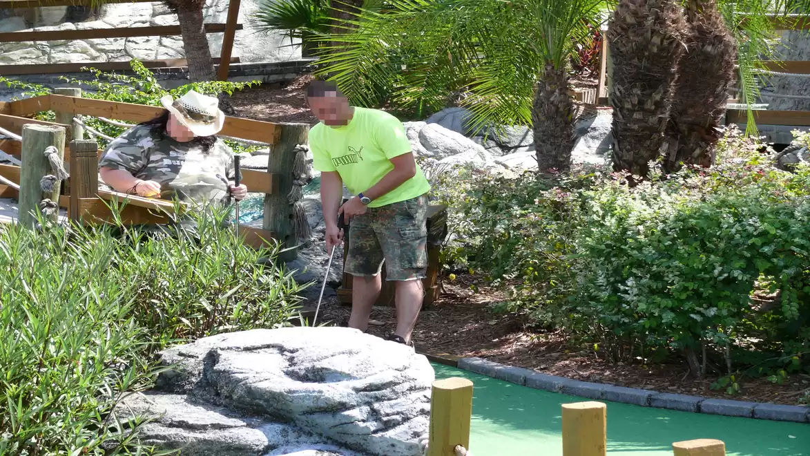 Two people are playing mini golf in a lush, green environment surrounded by plants. One person is wearing a light green shirt and is focused on the shot as the other watches.