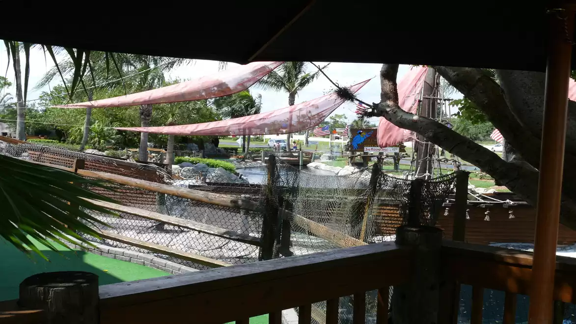 An outdoor area with palm trees, a wooden fence, and partially covered with red sunshades. In the background, various structures suggest an amusement or adventure park.
