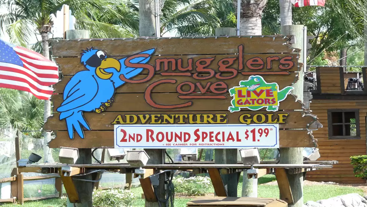 A large wooden sign displays the 'Smugglers Cove Adventure Golf' attraction featuring a blue parrot and a sign for a second round at $1.99. Palm trees and an American flag are visible in the background.