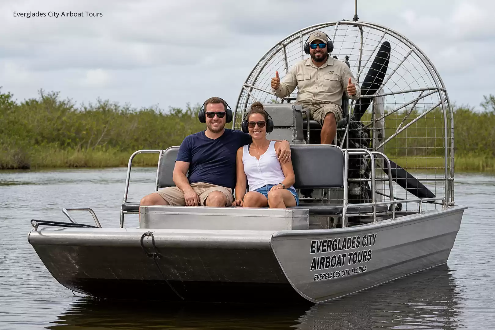 Three people sitting on an airboat in the Everglades, surrounded by water and vegetation.