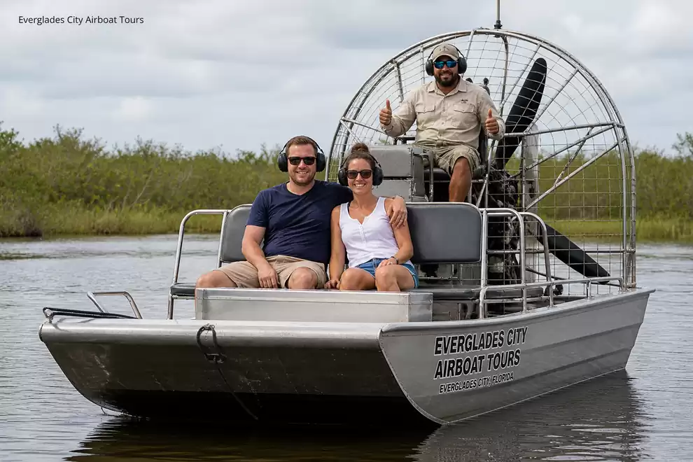 Three people sitting on an airboat in the Everglades, surrounded by water and vegetation.