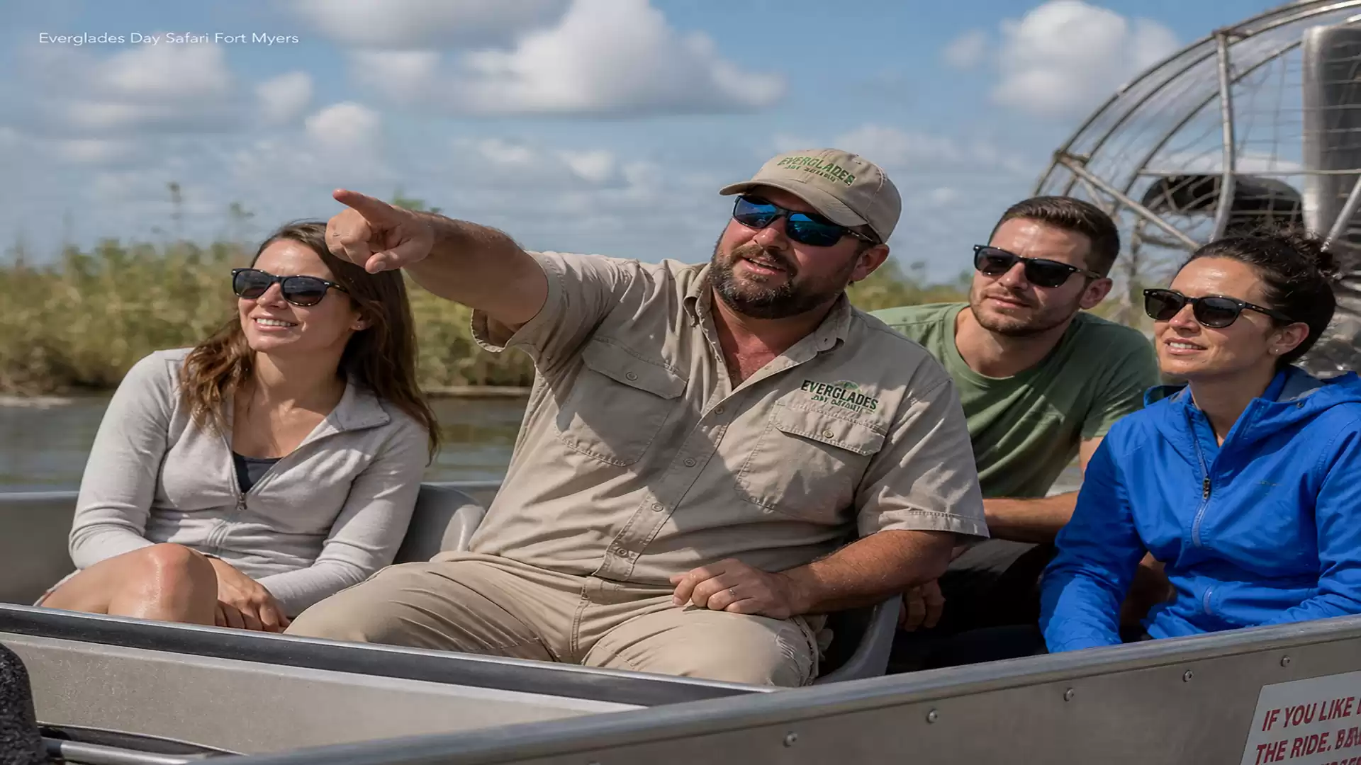 Four people on an airboat tour in a swamp area on a sunny day. The guide is pointing at something in the distance.