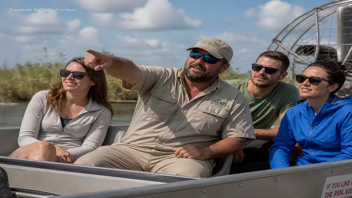 Four people on an airboat tour in a swamp area on a sunny day. The guide is pointing at something in the distance.