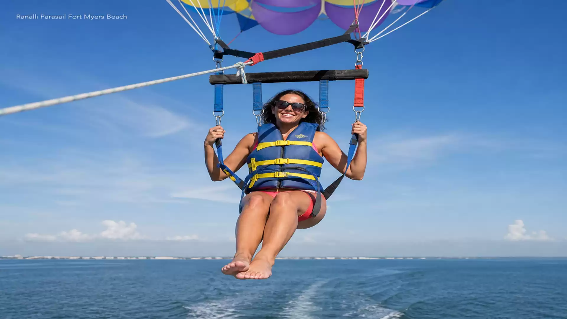 A woman enjoys parasailing with a colorful parachute over the open sea under a bright blue sky.