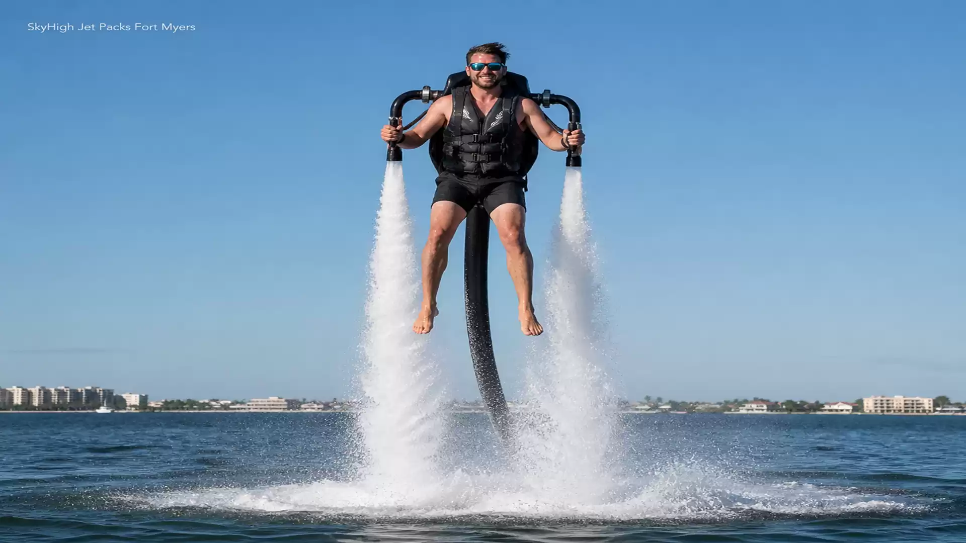 A man wearing sunglasses is flying with a water jetpack over the water on a sunny day.