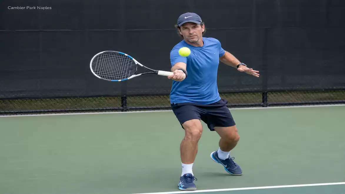 A man in a blue shirt playing tennis on a hard court. He hits the ball with his tennis racket.