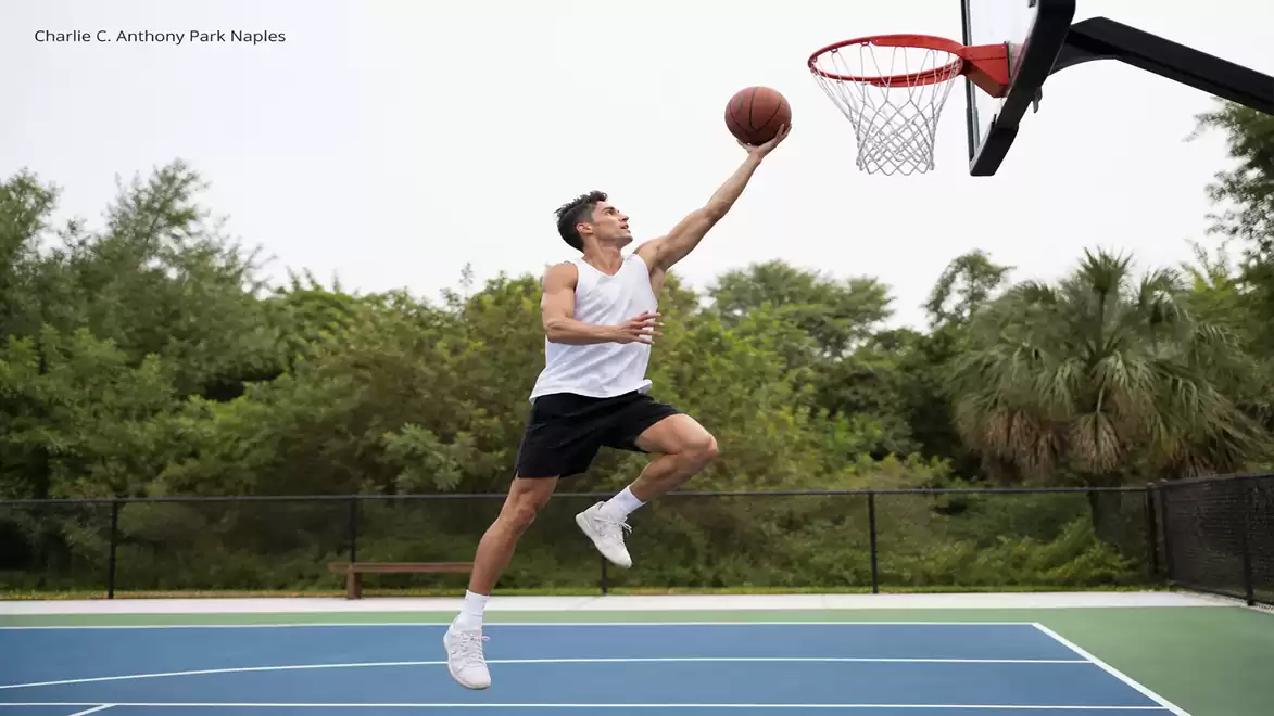 A basketball player in a white jersey and black shorts jumps to shoot a ball into the hoop on an outdoor court surrounded by trees.