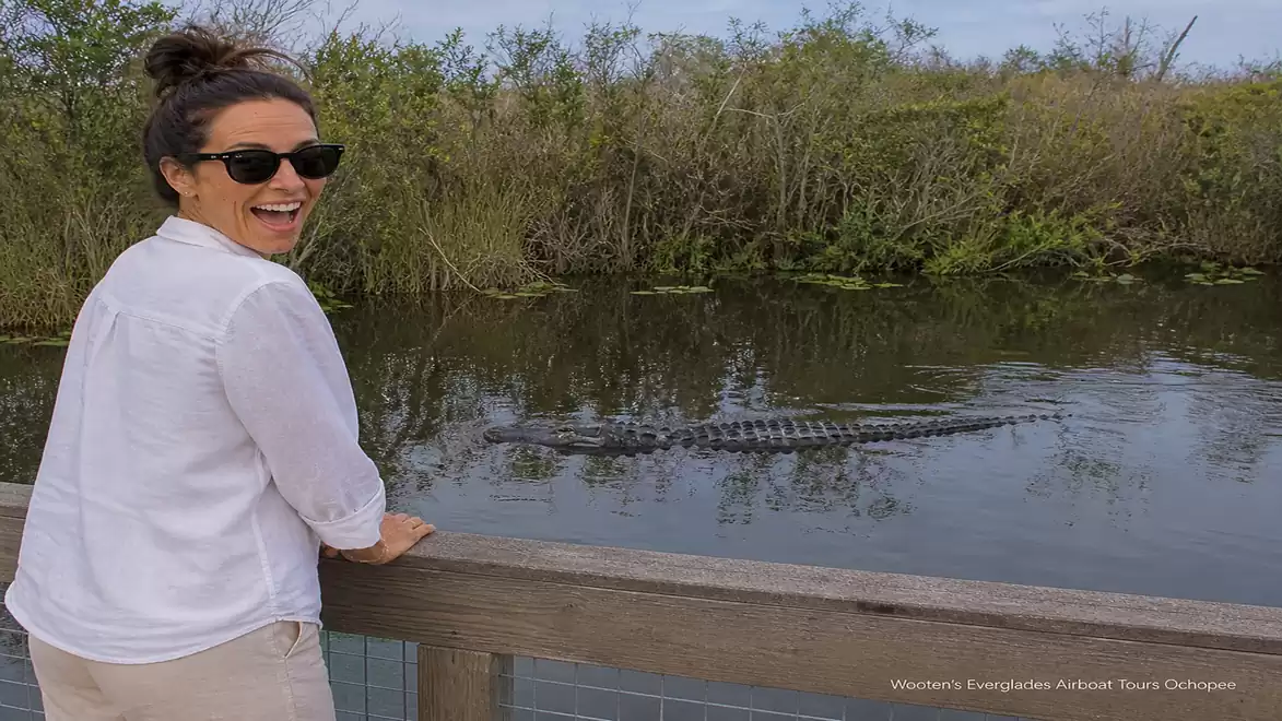 A smiling woman with sunglasses stands by a wooden fence, observing an alligator in the water in a nature park.