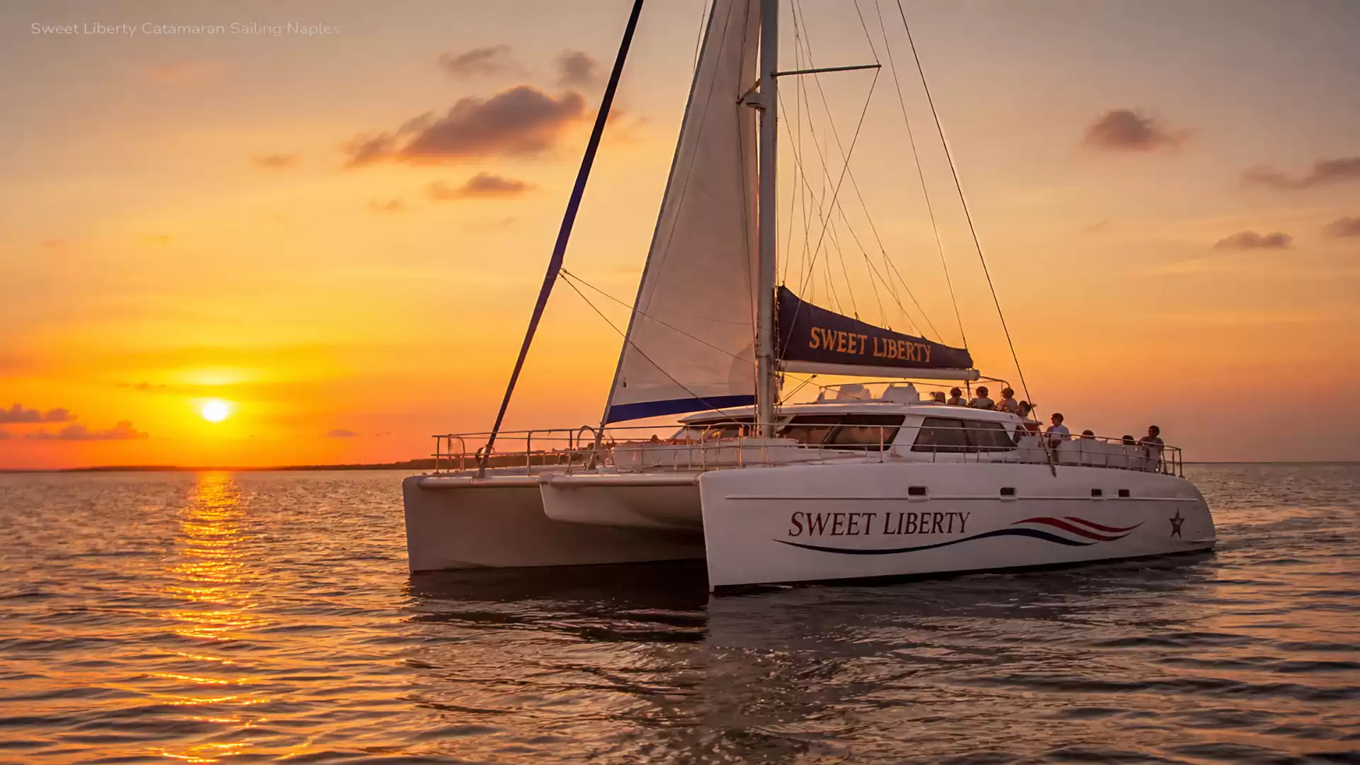 A catamaran sails on the sea at sunset in warm orange and gold tones.