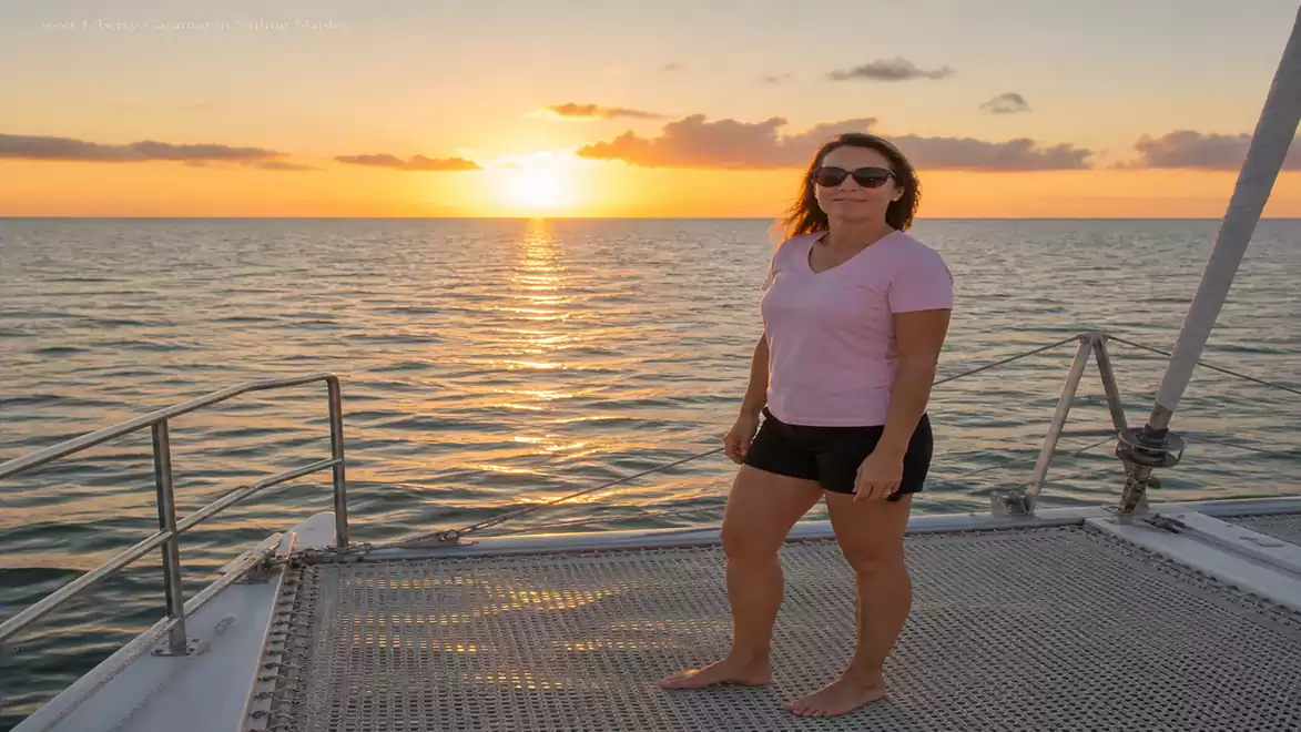 A woman stands on a boat deck in front of a sunset over the sea.
