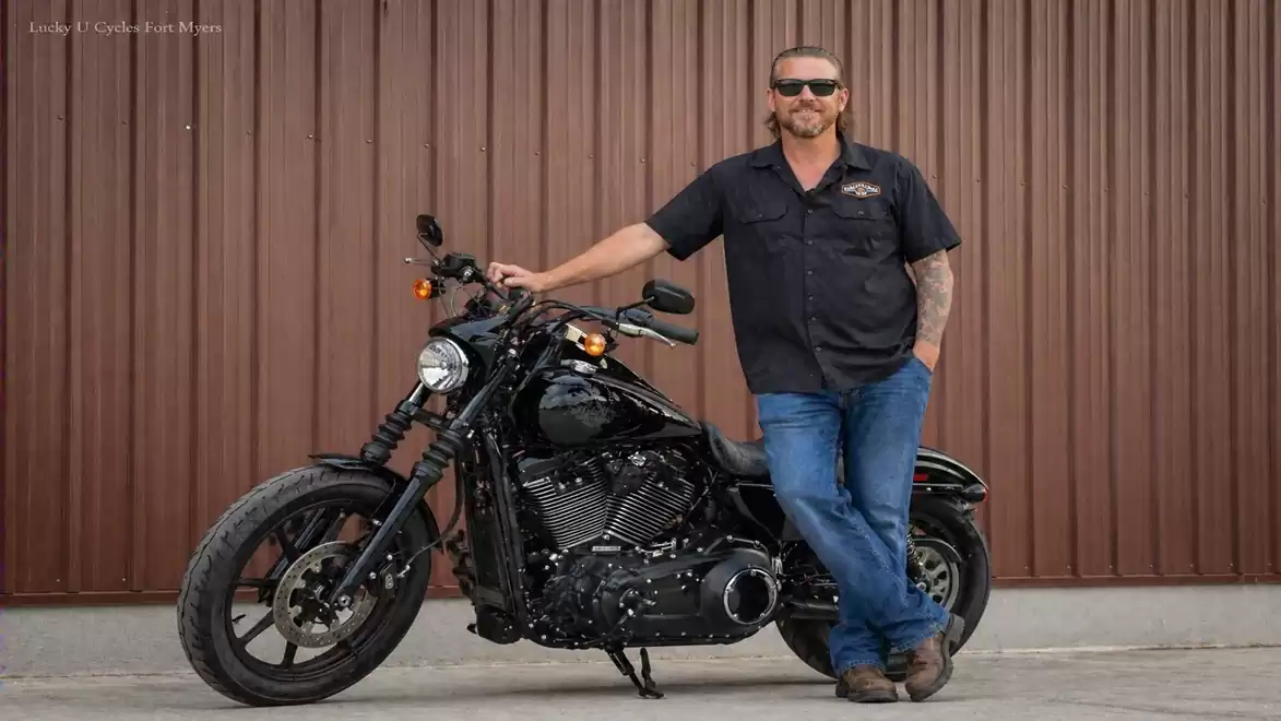 Man leaning on a black motorcycle in front of a brown metal wall.