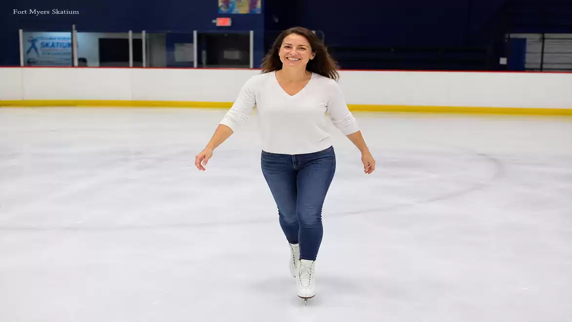 A woman in a white top and blue jeans happily ice skating on an ice rink.