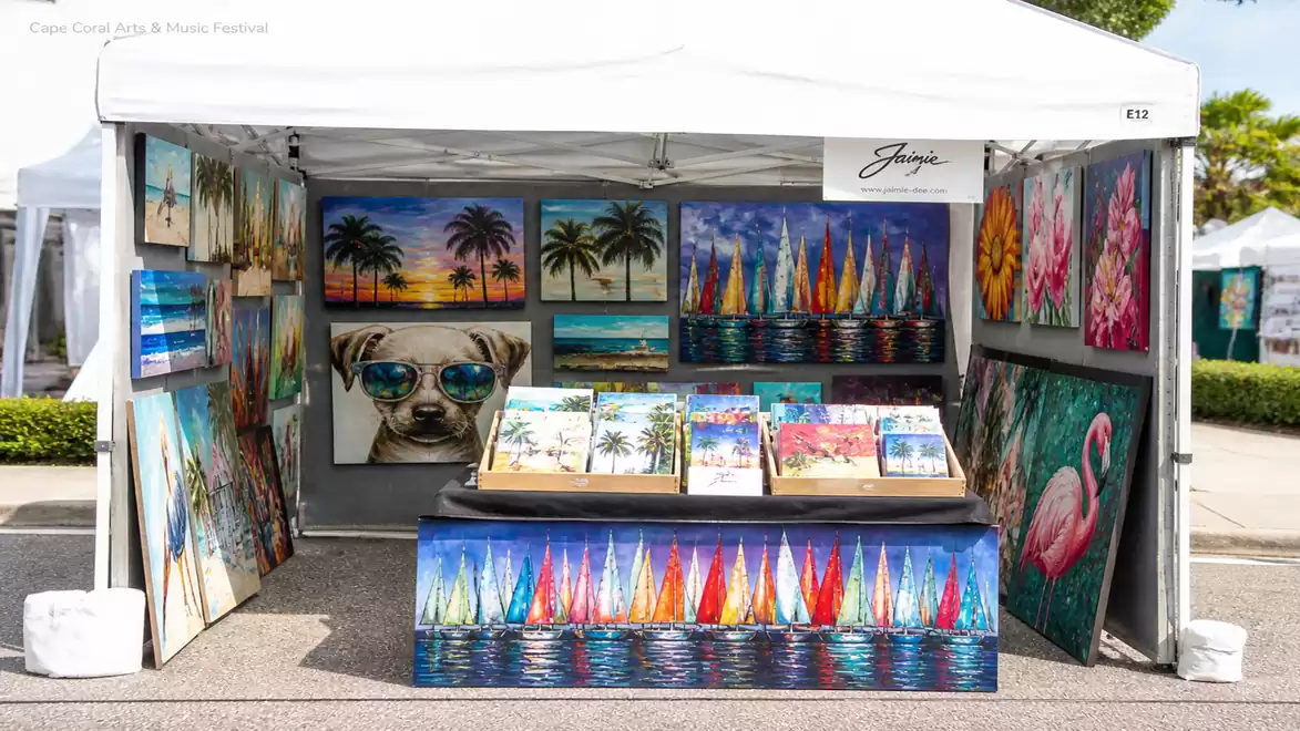 Art booth with brightly colored paintings of sailboats, palm trees, and animals at an outdoor exhibition.