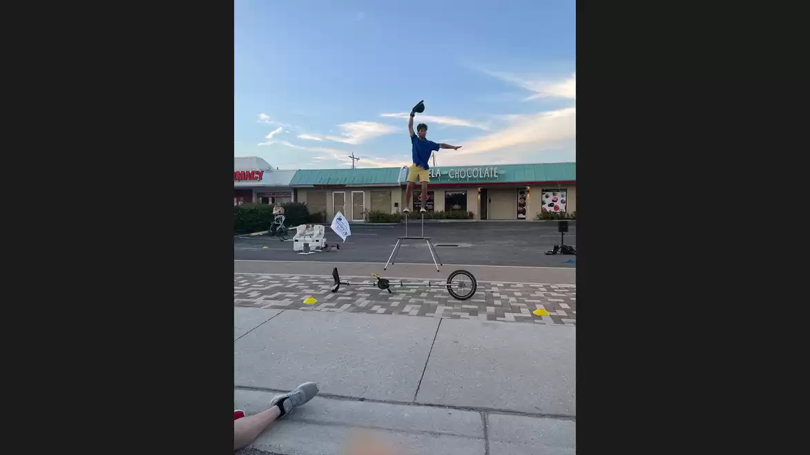 A man balances on a tall unicycle on a busy street, with shops visible in the background.