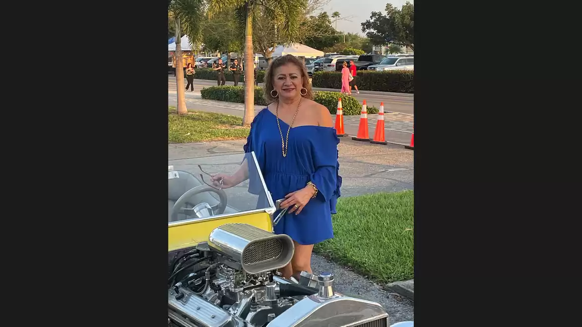 Woman in a blue dress stands next to a motorcycle on the street with trees and traffic cones in the background.