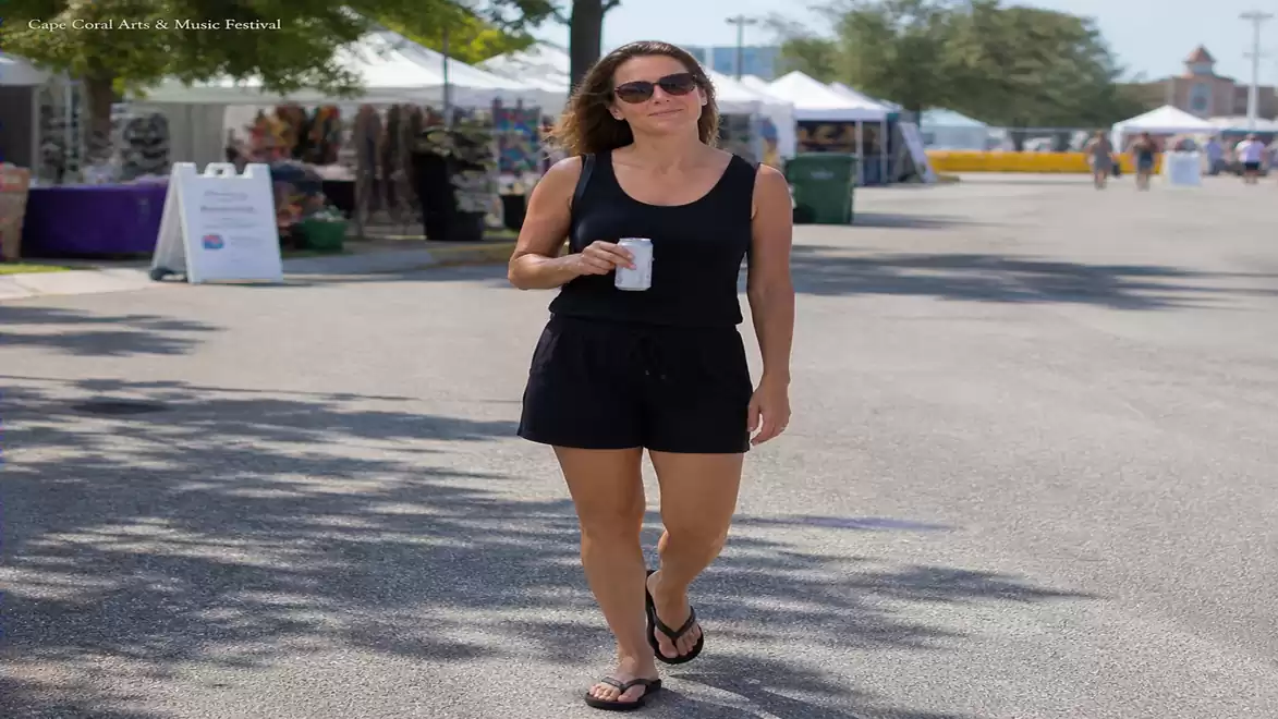 A woman in black clothing walking with a drink in hand at an outdoor festival, surrounded by trees and booths.