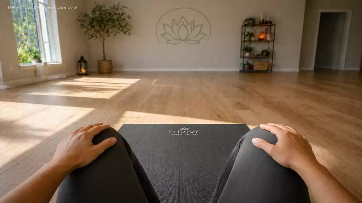 Inside view of a serene yoga studio with a perspective from a mat, hands in foreground, bright room, and plants in the corner.