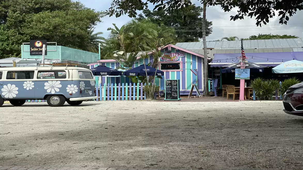 A colorful café with retro charm and a hippie-style VW bus in the foreground on a sunny parking lot.