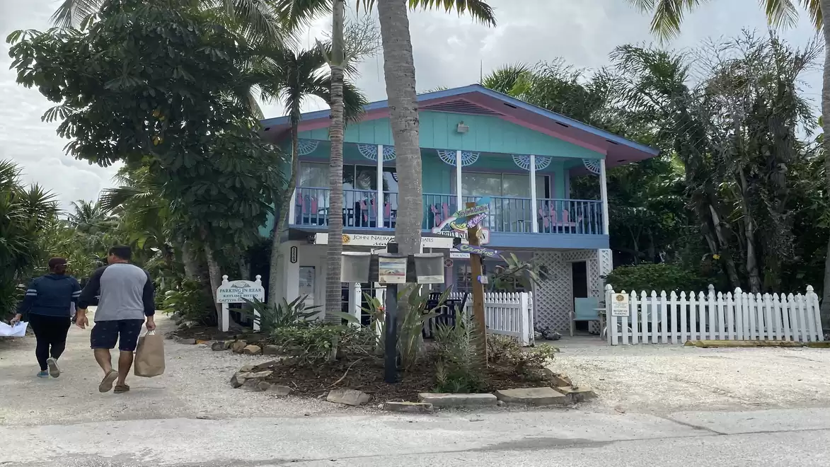 A colorful two-story house with blue accents on a tropical island. There are palm trees and a small white fence in front of the house. Two people are walking with bags on the left side of the image.