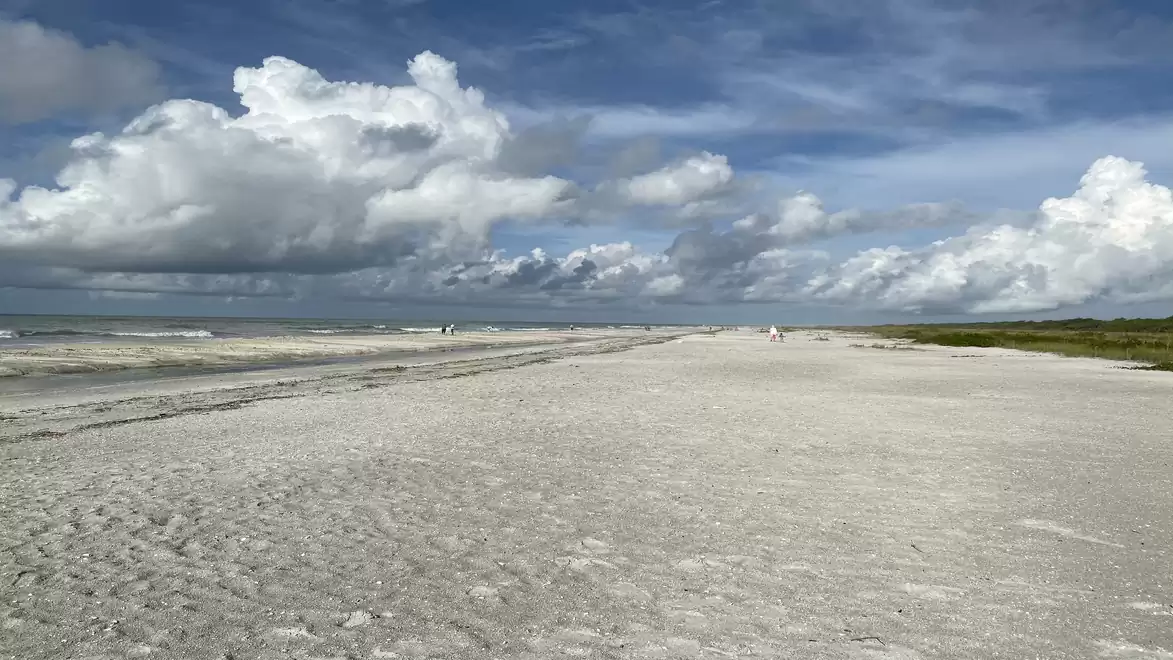 A wide, deserted sandy beach under a cloudy sky with some clouds on the horizon.
