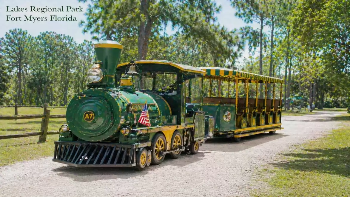 A green train in Lakes Regional Park, Fort Myers, Florida, travels along a narrow gravel road, surrounded by trees and green areas.