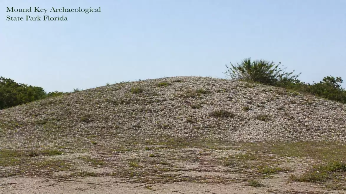 A large, grassy mound in Mound Key Archaeological State Park in Florida, surrounded by trees under a blue sky.