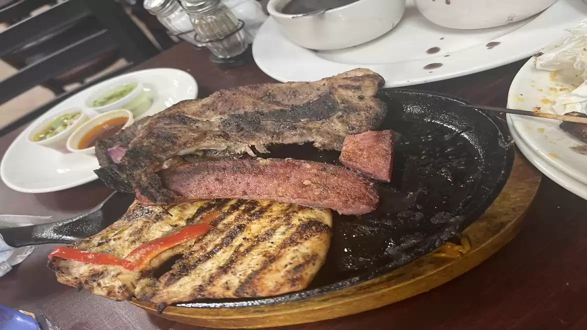Close-up of grilled meat served on a wooden board, accompanied by sauces in small bowls on a table. Plates with leftovers are visible in the background.