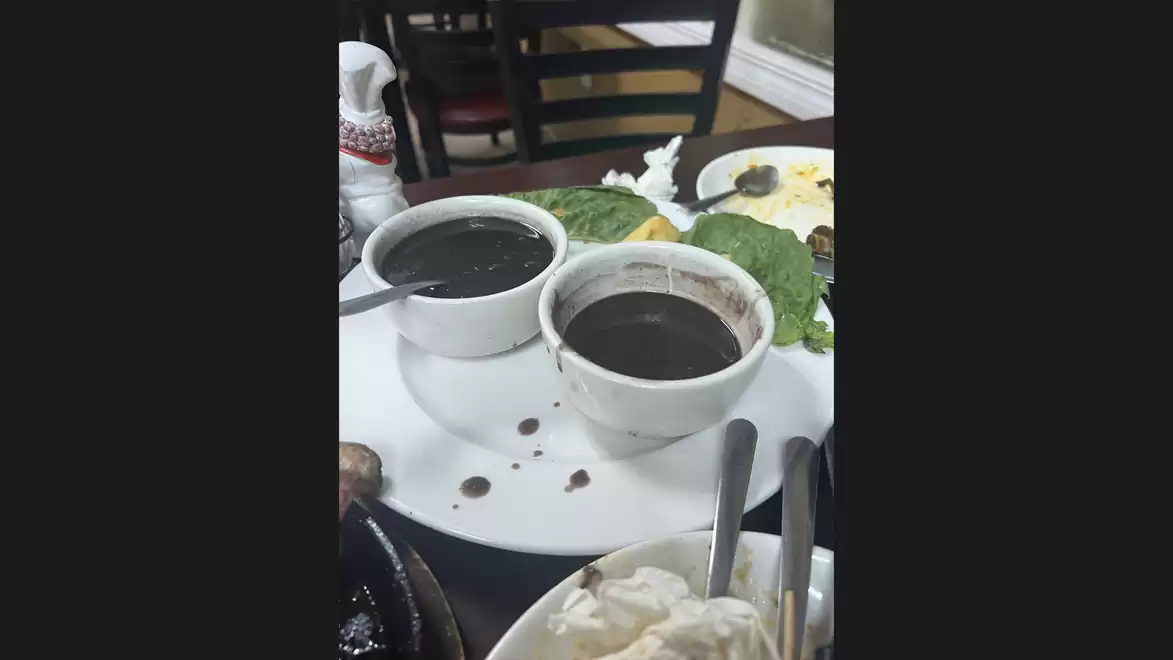 A restaurant table with several dishes, two bowls filled with a dark soup or sauce, and lettuce leaves. Chairs and additional dishware are visible in the background.
