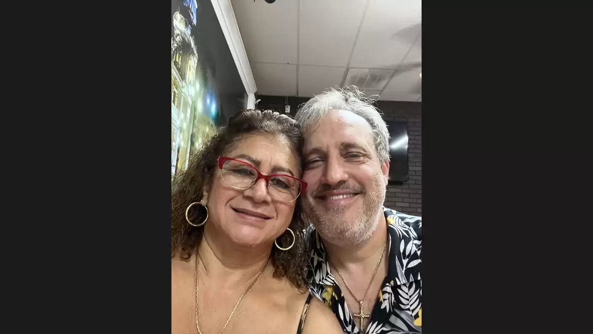 A smiling couple posing indoors. Both are wearing casual clothing and standing close together.
