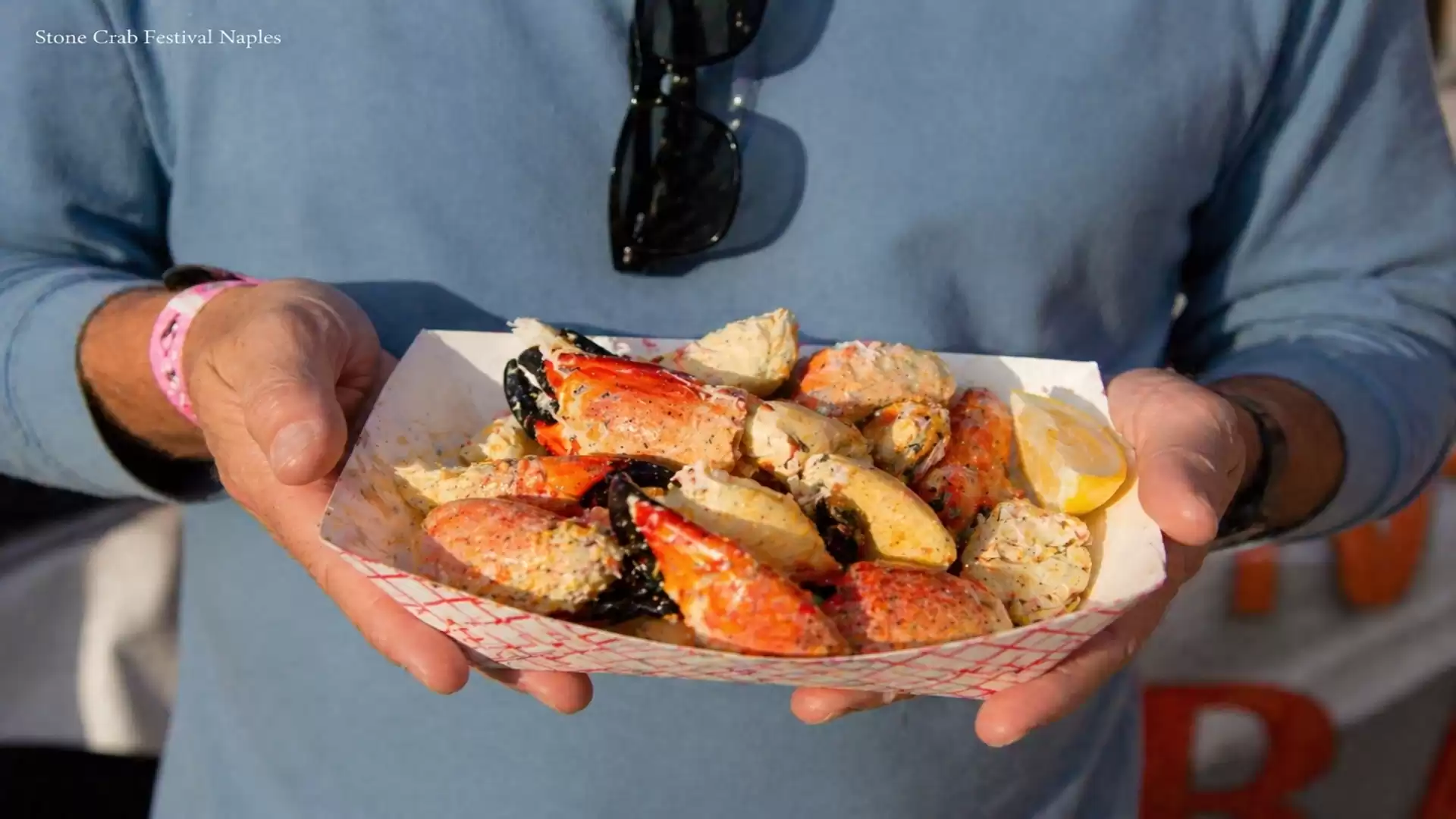 A man holds a tray of freshly prepared crab legs garnished with lemon slices against a blurred background at the Stone Crab Festival in Naples.