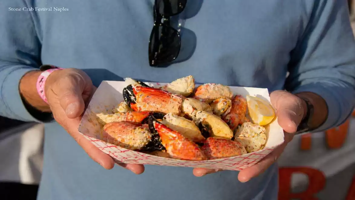 A man holds a tray of freshly prepared crab legs garnished with lemon slices against a blurred background at the Stone Crab Festival in Naples.