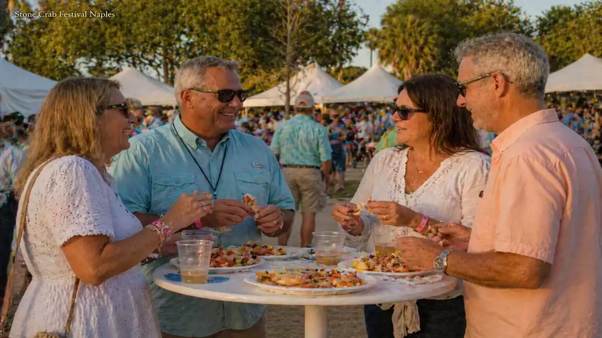 Four people stand around a table outdoors, eating pizza and laughing. Trees and tents are visible in the background, suggesting a festival or celebration.