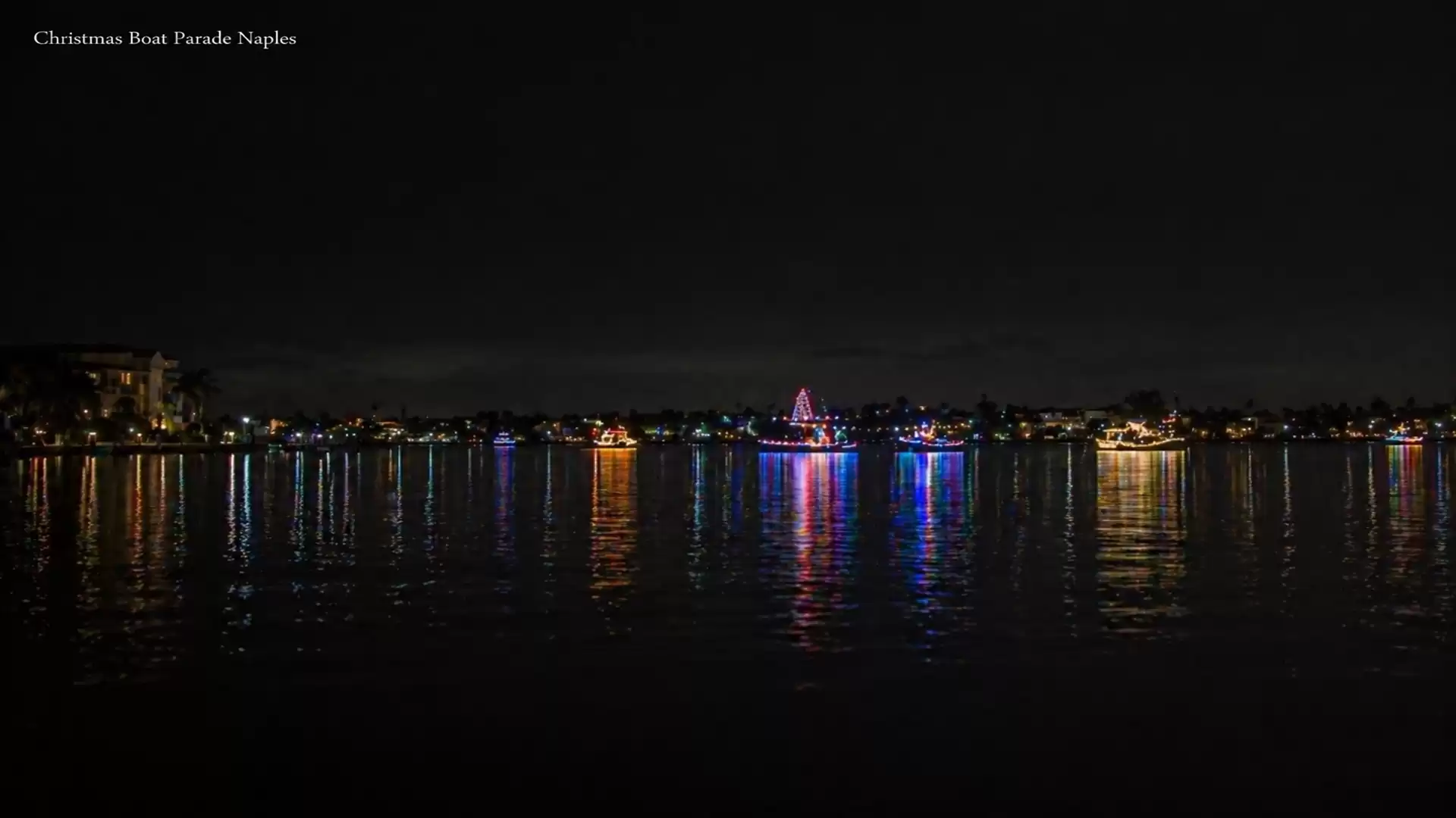 Night view of a Christmas light boat parade on water, with lights reflecting in the water.