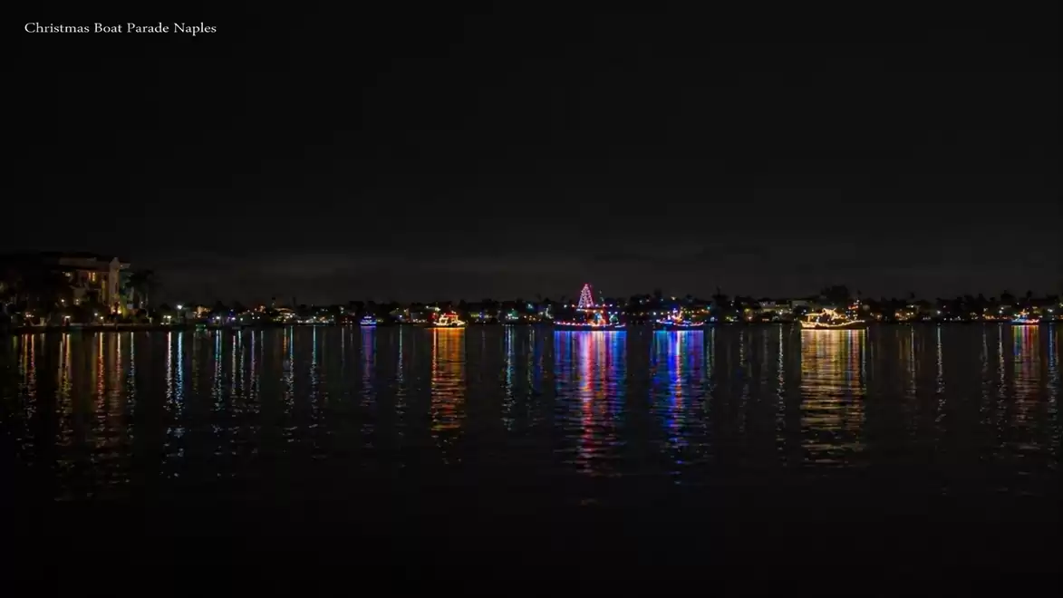 Night view of a Christmas light boat parade on water, with lights reflecting in the water.