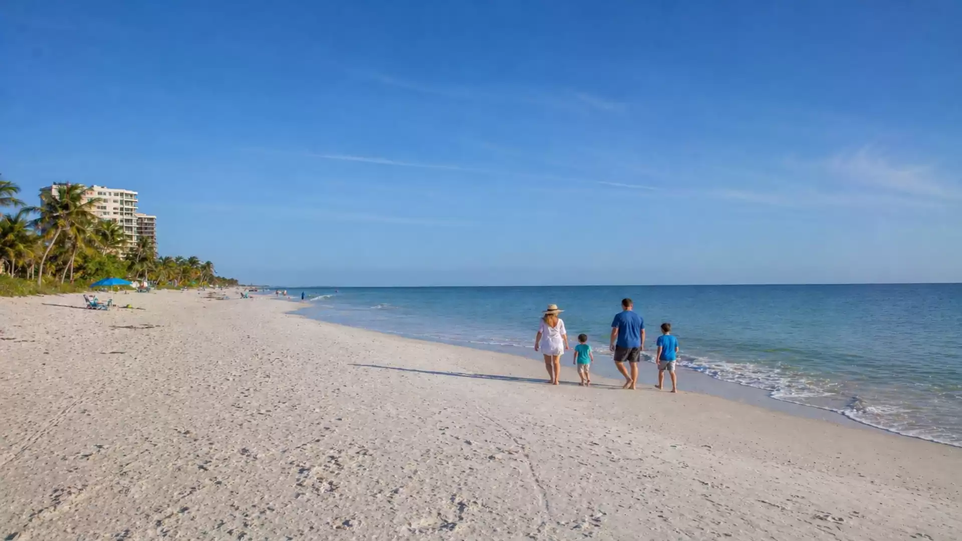 A couple with two children walks along a sunny sandy beach, with palm trees and buildings visible in the background.