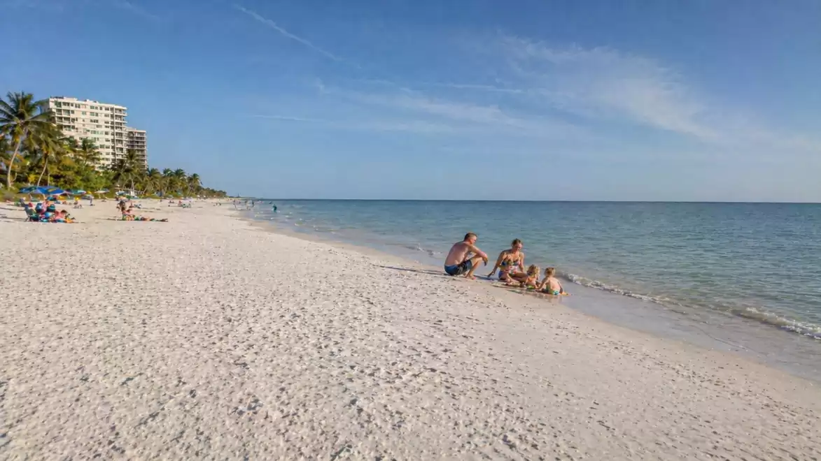 A sunny beach with some people enjoying the sea. Palm trees and a hotel building are visible in the background.