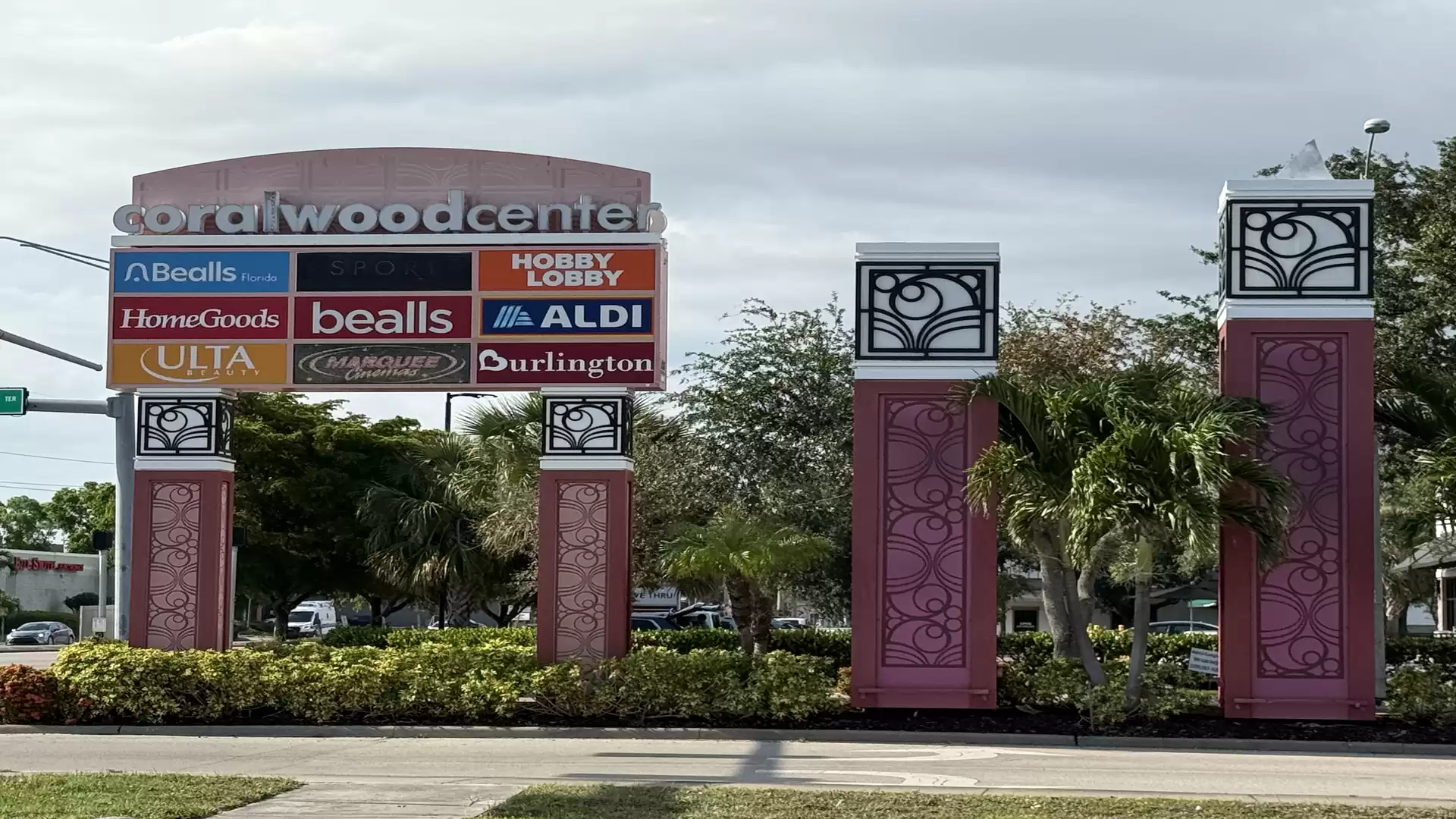 Entrance area of the Coralwood Center with several store signs from brands like Bealls, Hobby Lobby, and Aldi. Surrounded by lush vegetation and a paved road in the foreground.
