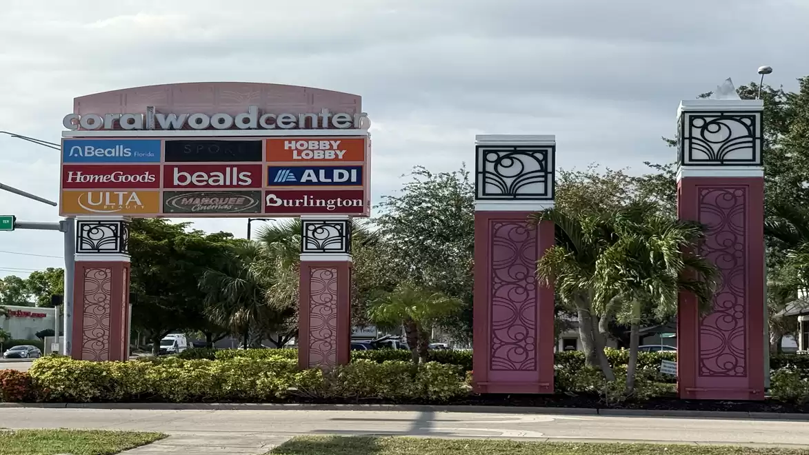 Entrance area of the Coralwood Center with several store signs from brands like Bealls, Hobby Lobby, and Aldi. Surrounded by lush vegetation and a paved road in the foreground.